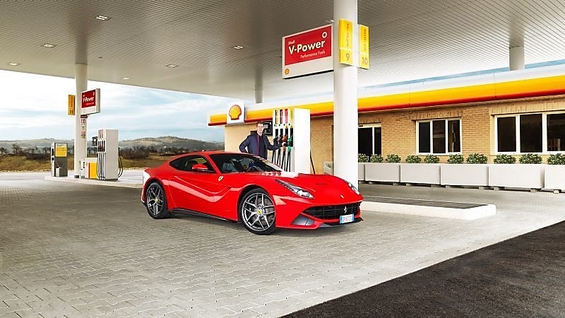 A red Ferrari sitting on a Shell station forecourt with a man leaning on a petrol pump